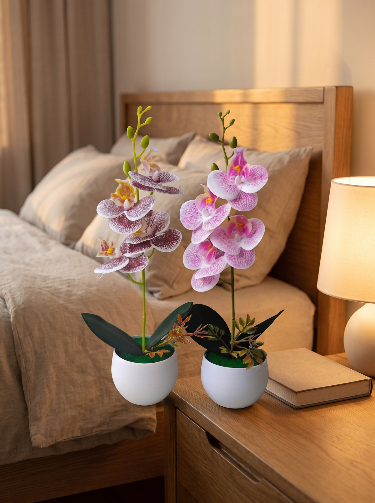 Faux orchid in a pot on a bedside table beside a book and a lamp